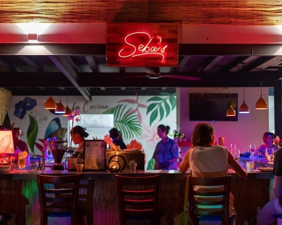 Seba's bar interior with neon sign, tropical decor, and patrons at the counter